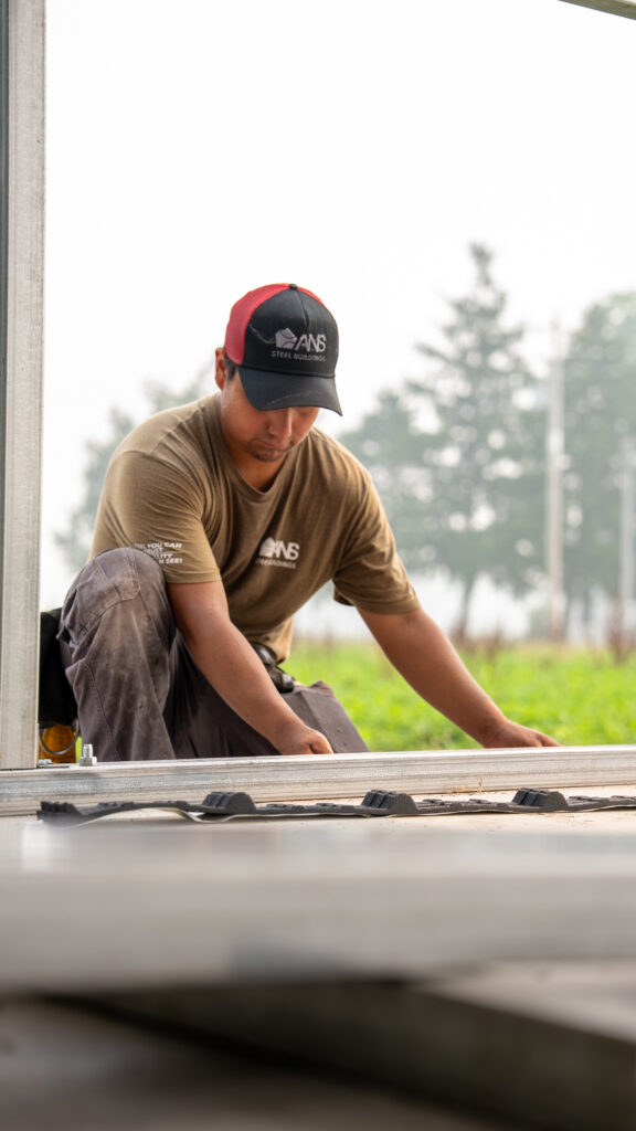A worker wearing ANS Steel Buildings clothing works beside the base of a steel building frame.