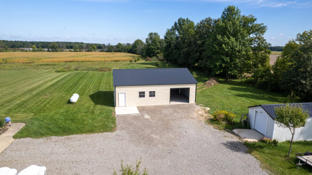 Tan metal garage with a dark roof, two windows, a white entry door, and an open garage bay beside a gravel driveway.